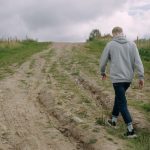 Two individuals walking on a rural dirt path surrounded by grass under a cloudy sky, creating a serene outdoor scene.