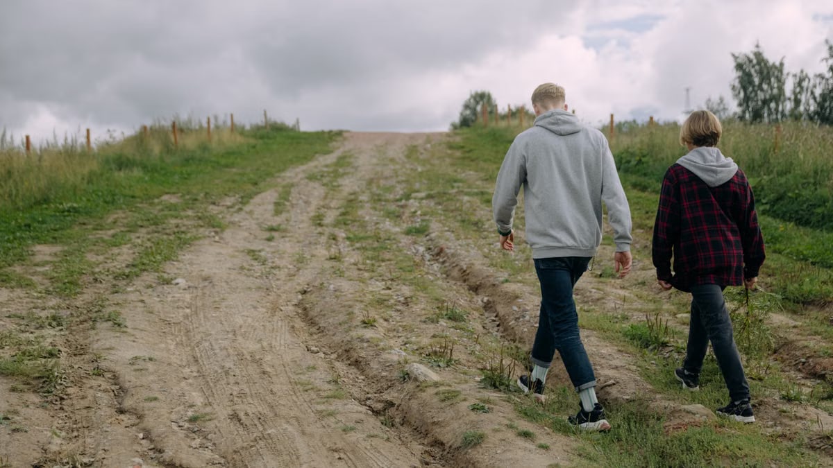 Two individuals walking on a rural dirt path surrounded by grass under a cloudy sky, creating a serene outdoor scene.